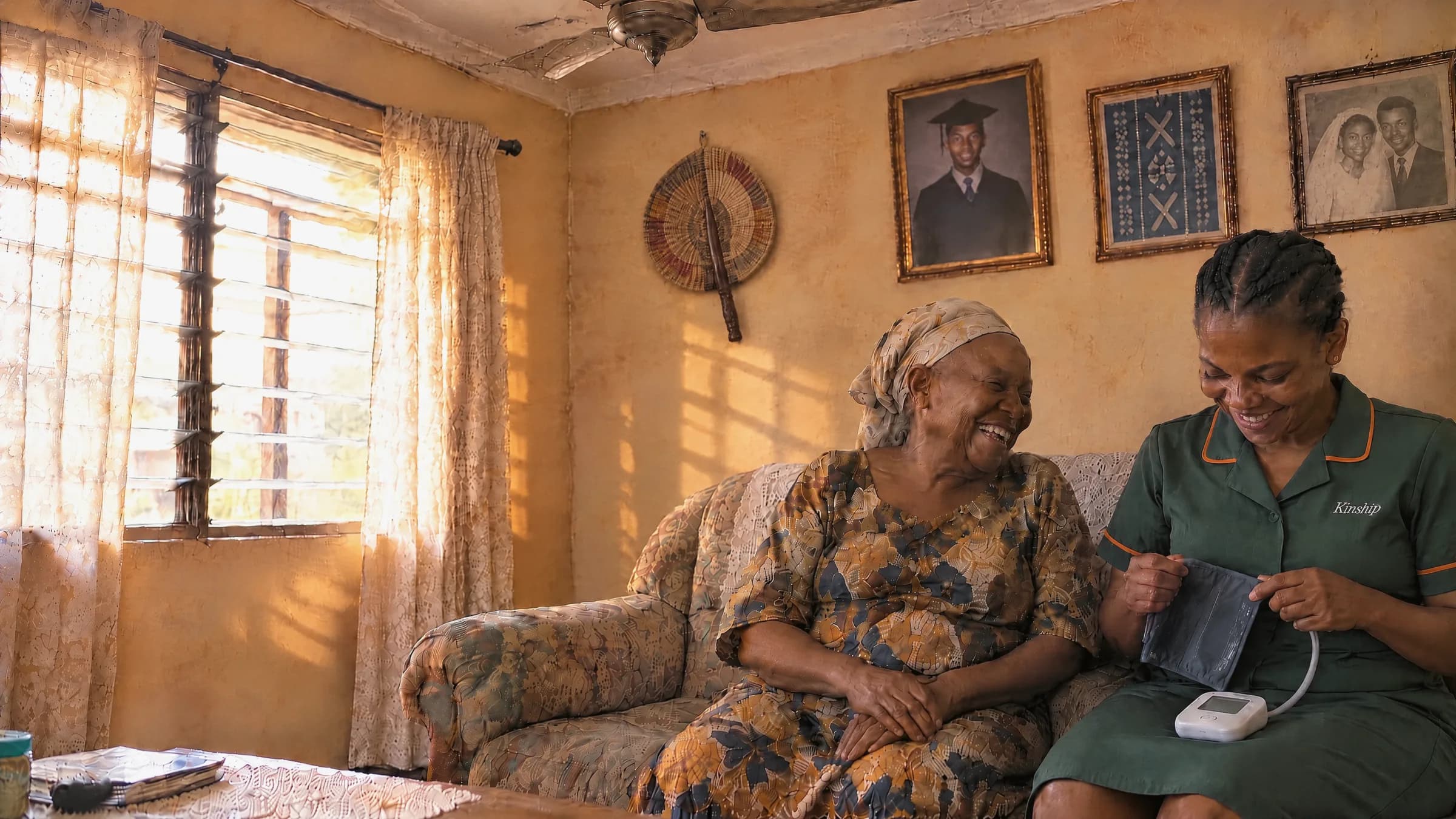 An older woman laughing on her sofa at home in Lagos as a Kinship nurse beside her unpacks a blood pressure cuff, in the afternoon sun